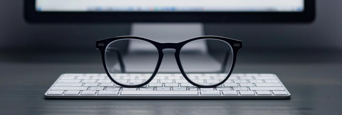 Une paire de lunettes noires repose au-dessus d'un clavier d'ordinateur blanc avec un moniteur d'informatique flou en arrière-plan.